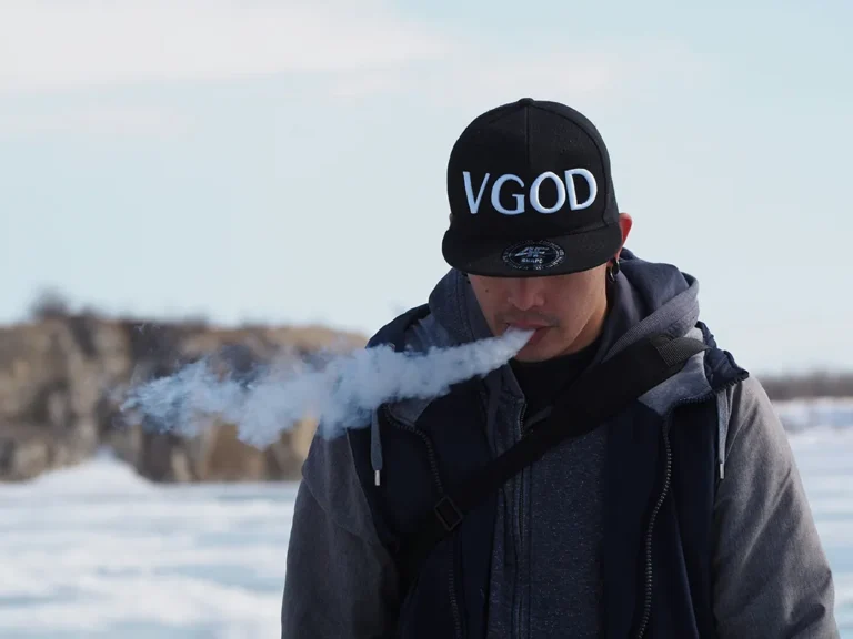 一位戴著「VGOD」品牌帽子和灰色連帽衫的成年男性老玩家,在模糊的雪地戶外冬日場景中專注地吞吐厚實的白色電子煙霧化蒸汽。這是一張用於現代替煙方案指南的特色圖片。